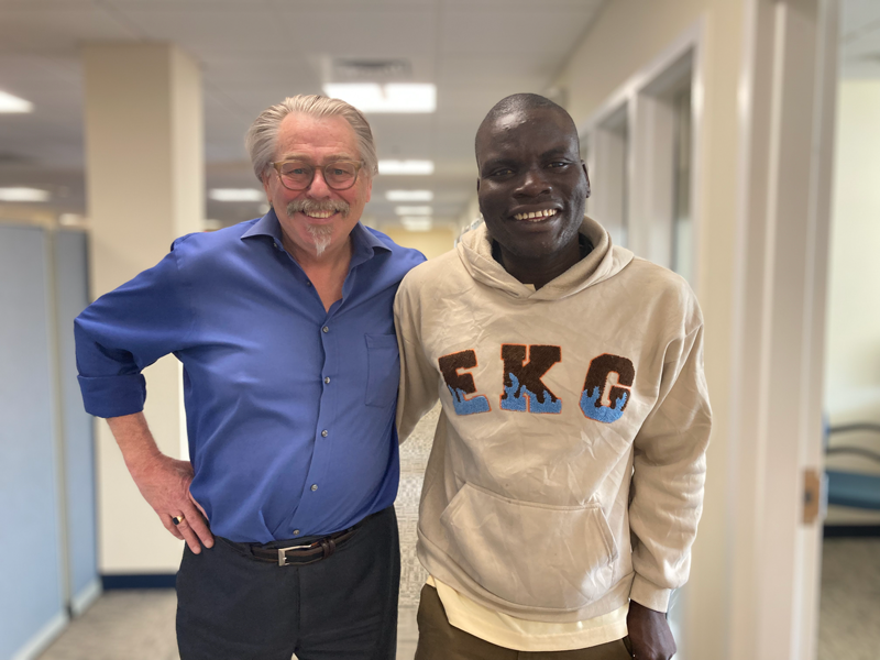 Two individuals, a white American man and a Black Ugandan man posing together in a hallway