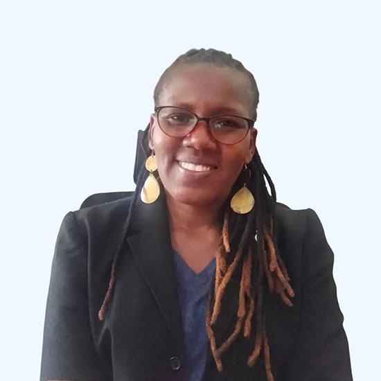 An African woman with long braided hair, wearing glasses, big gold earrings, and a blazer, smiles at the camera.