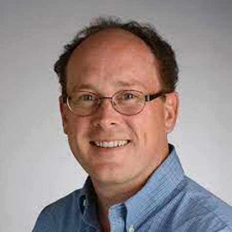 Photo of a white American man with brown hair, wearing eyeglasses and a light blue shirt, smiling at the camera.