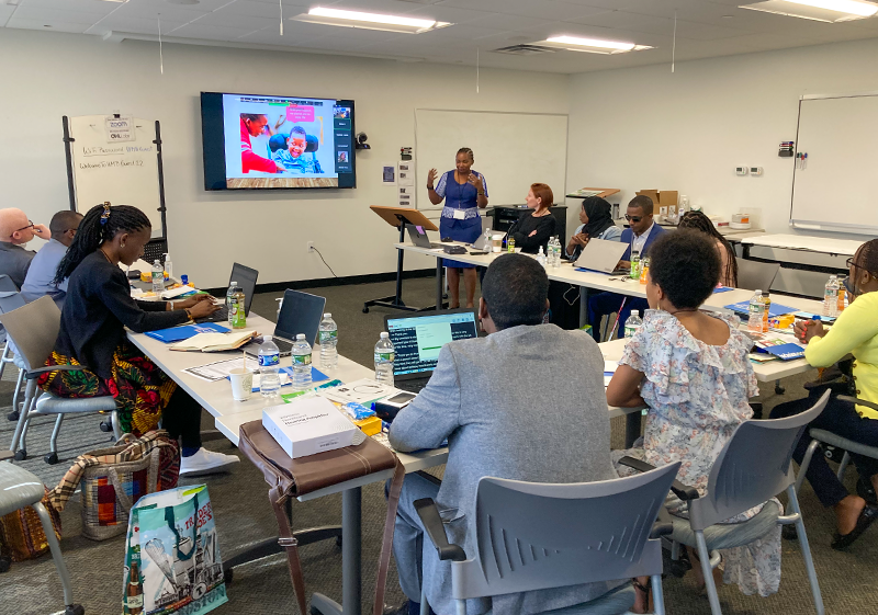 A group of 13 people are in a conference room with a woman at the front giving a presentation.