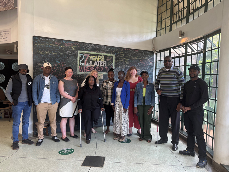 Eleven individuals pose for a group photo in front of a black board that reads, '27 years later and where were you?'