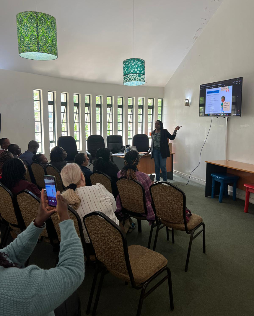 A woman stands at the front of a meeting room, delivering a presentation while slides are projected on a screen and participants are seated and listening.