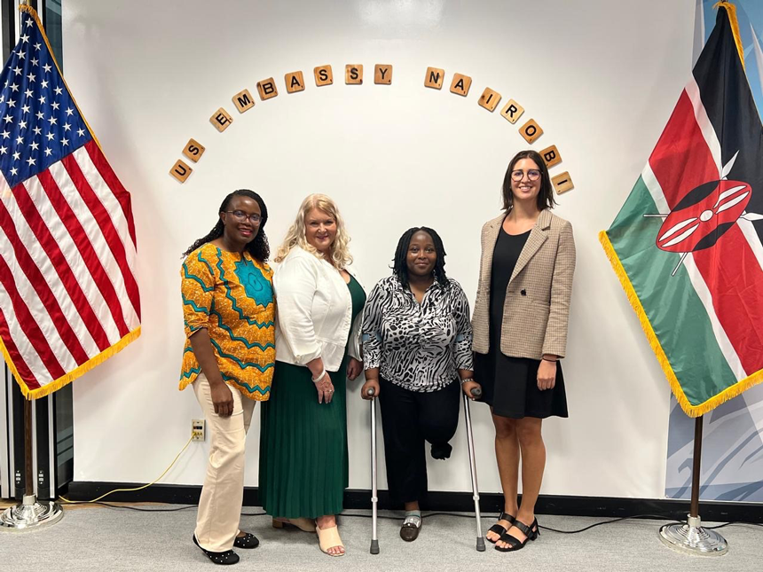 Four women smile while posing for a group photo. The US flag stands to their left and the Kenyan flag to their right. Above them is a sign reading 'US Embassy Nairobi.' 