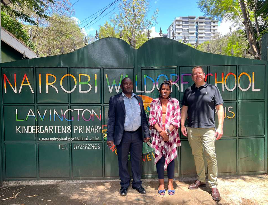 Three individuals—two men and one woman—pose for a group photo in front of a green gate bearing the inscription 'Nairobi Waldorf School.'