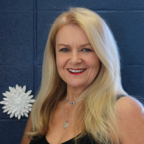 A woman with long blonde hair, wearing a sleeveless blouse, smiling at the camera.