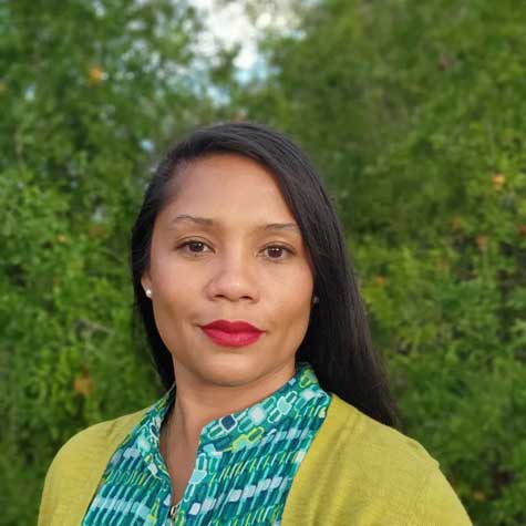 Photo of a woman with long black hair, dressed in a green blouse and cardigan, against a backdrop of trees, looking directly into the camera.