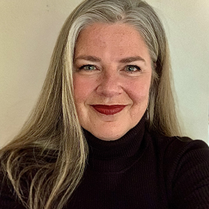 Profile of a white woman with long blond and gray hair, wearing a black turtleneck, smiling into the camera.