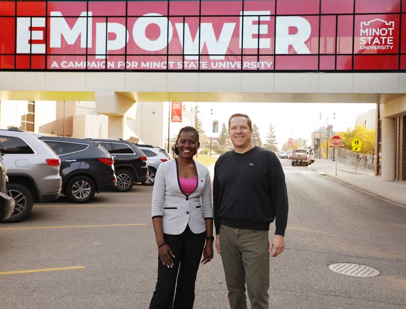 Two individuals, a Black Ugandan woman and white American man, posing in a car park with a covered walkway in the background. On the walkways it says “EMPOWER – A campaign for Minot State University with the university’s logo next to it.