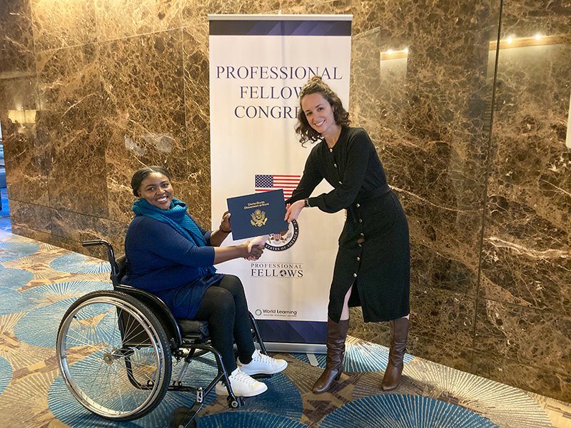 An African Fellow sitting in a wheelchair receiving a blue certificate of from a US program officer in a black dress. 