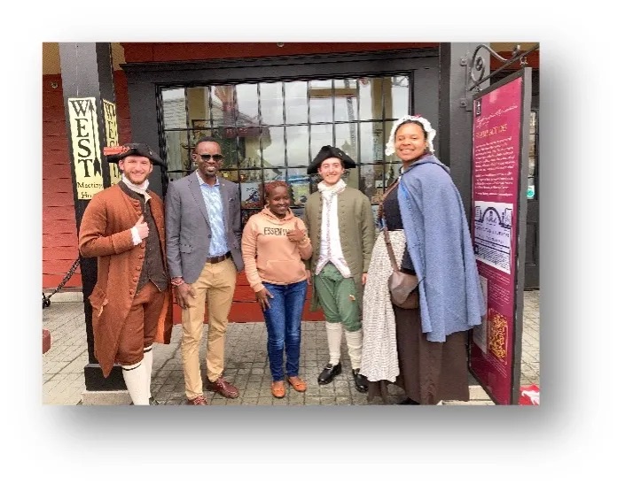 Two people wearing modern clothing (both African fellows) stand with three people where colonial clothing, (two white men and a Black woman).