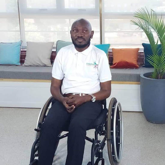 An African man with a beard is wearing a white shirt and black trousers. He is sitting in a wheelchair in a lounge area. 