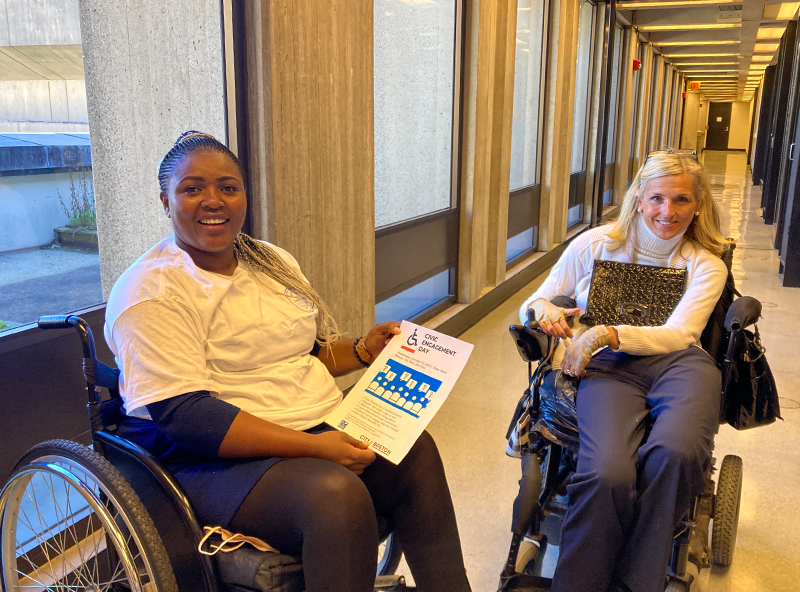 Two women in wheelchairs, one American and one African, posing in a hallway.