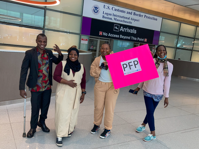 Four individuals, one man and three women, posing with a pink sign that reads 'PFP'. They are standing in an airport arrival area. 