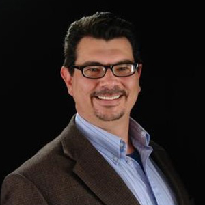 A profile picture of a white man with a short black beard, black hair, and glasses looks at the camera in front of a black background. 