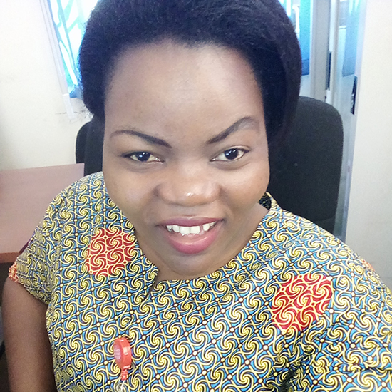 An African woman with short hair is wearing an African print blouse, taking a selfie while sitting in the office. 