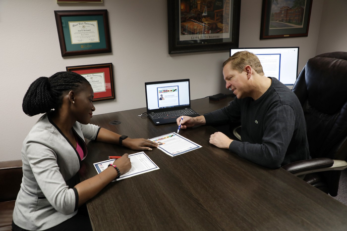 A Ugandan woman and a white man sit on either sides of a desk and review documents.