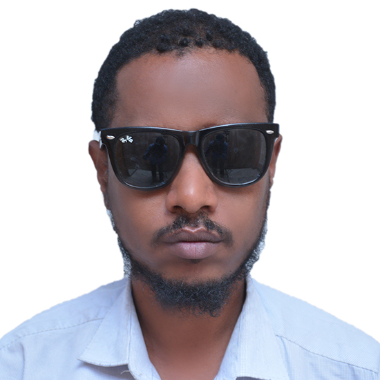 An African man with short black hair, a mustache, and a beard is wearing a light blue shirt and black sunglasses.