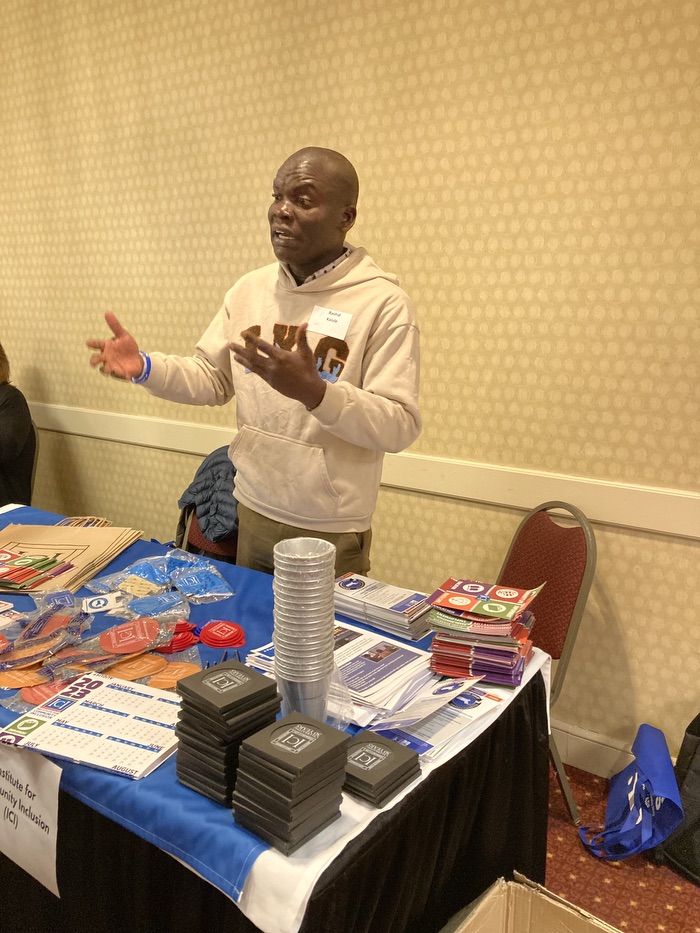An African man is standing behind a table filled with brochures, speaking to someone not pictured.