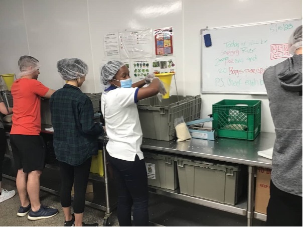 An African woman wearing a mask, gloves, and hair net is working beside three Americans on a metal worktable.