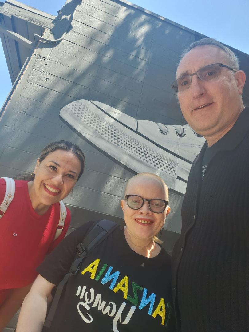 A tan woman with long, dark hearing, wearing a red shirt, a light-skinned woman with red hair and dark glasses, and a white, tall man with grey hair and black glasses stand together smiling at the camera.