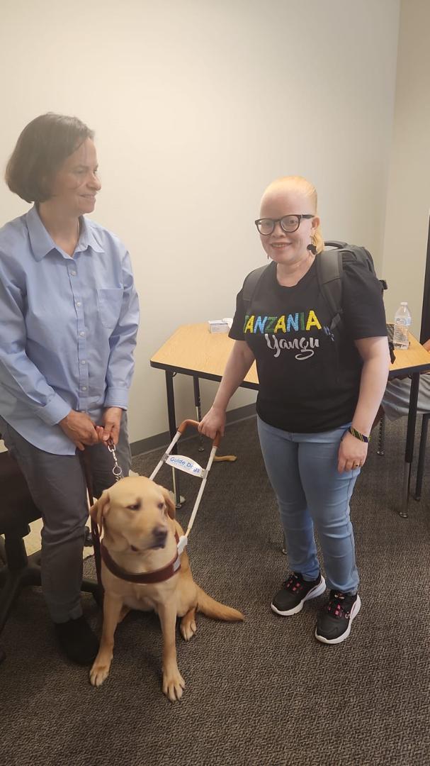 From left to right: A white woman with medium length brown hair, looking away from the camera, and holding the leash of a golden retriever dog. Standing beside her is a white, African woman wearing glasses and also holding onto the dog. 