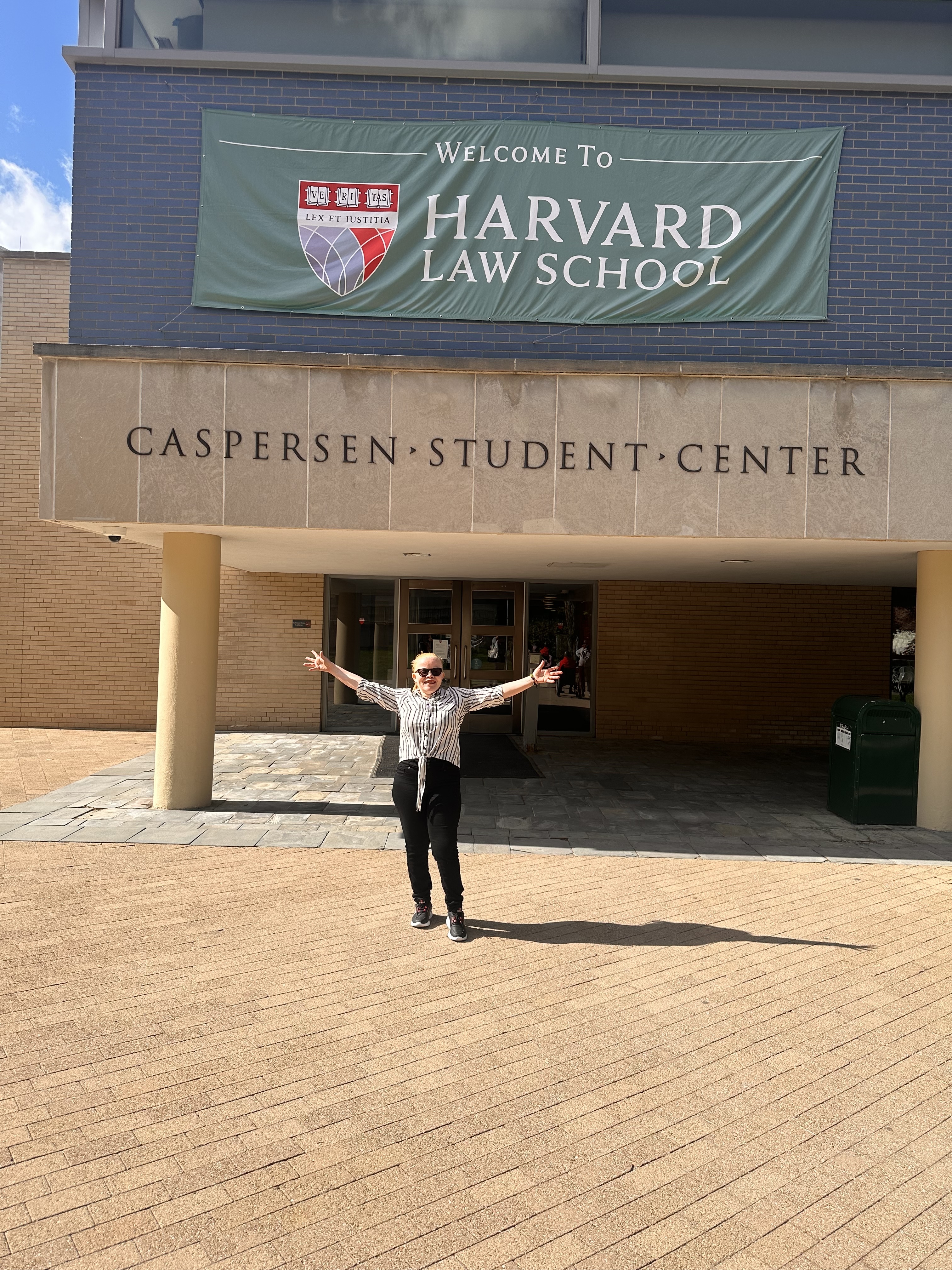 A light-skinned woman, with red hair and black glasses weaing a black and white polka dot shirt and black pants, stands with her arms outstretched under a sign that reads “Harvard Law School.”