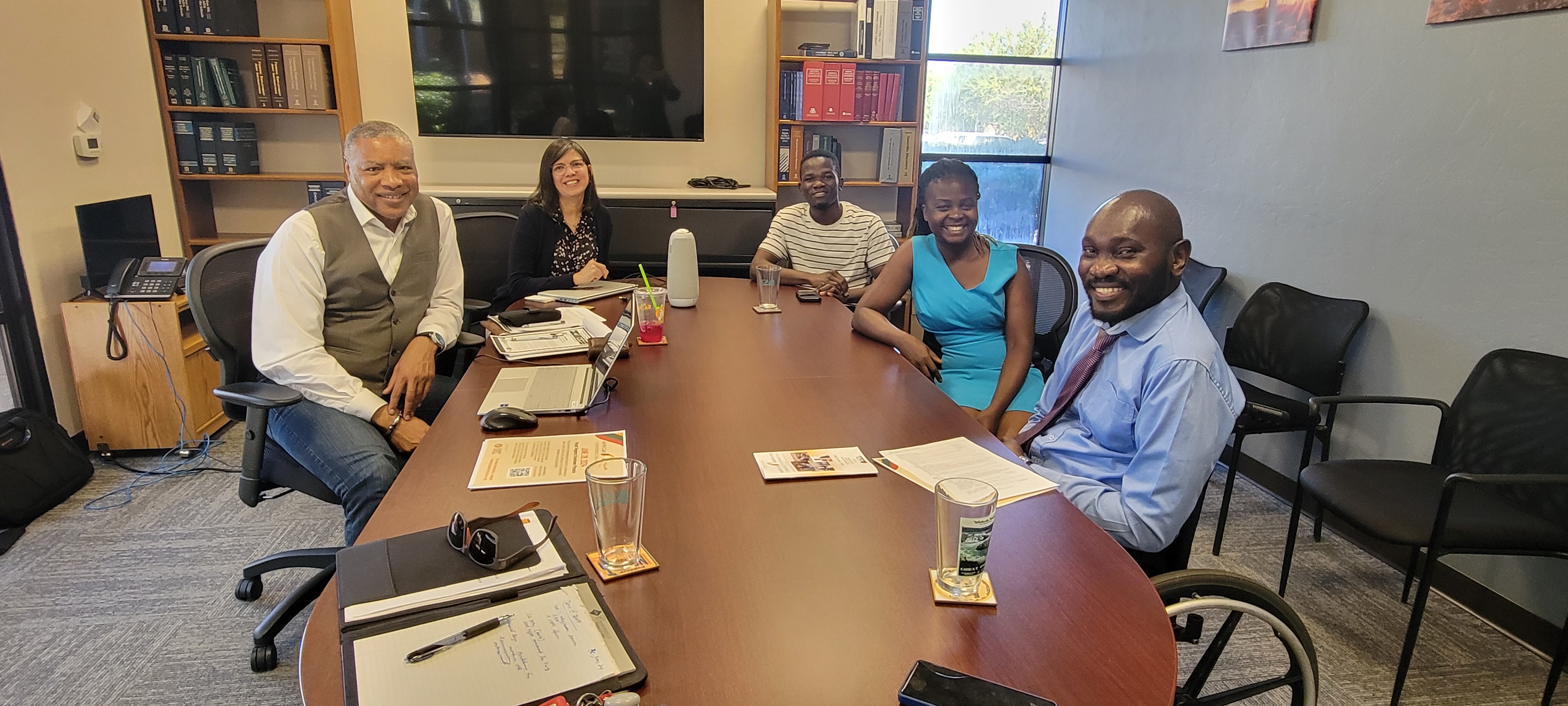 A group of five people, three men and two women, sitting at a table in a conference room, smiling for the camera. On the table are materials, cups with water, and in the background are bookshelves and chairs.  