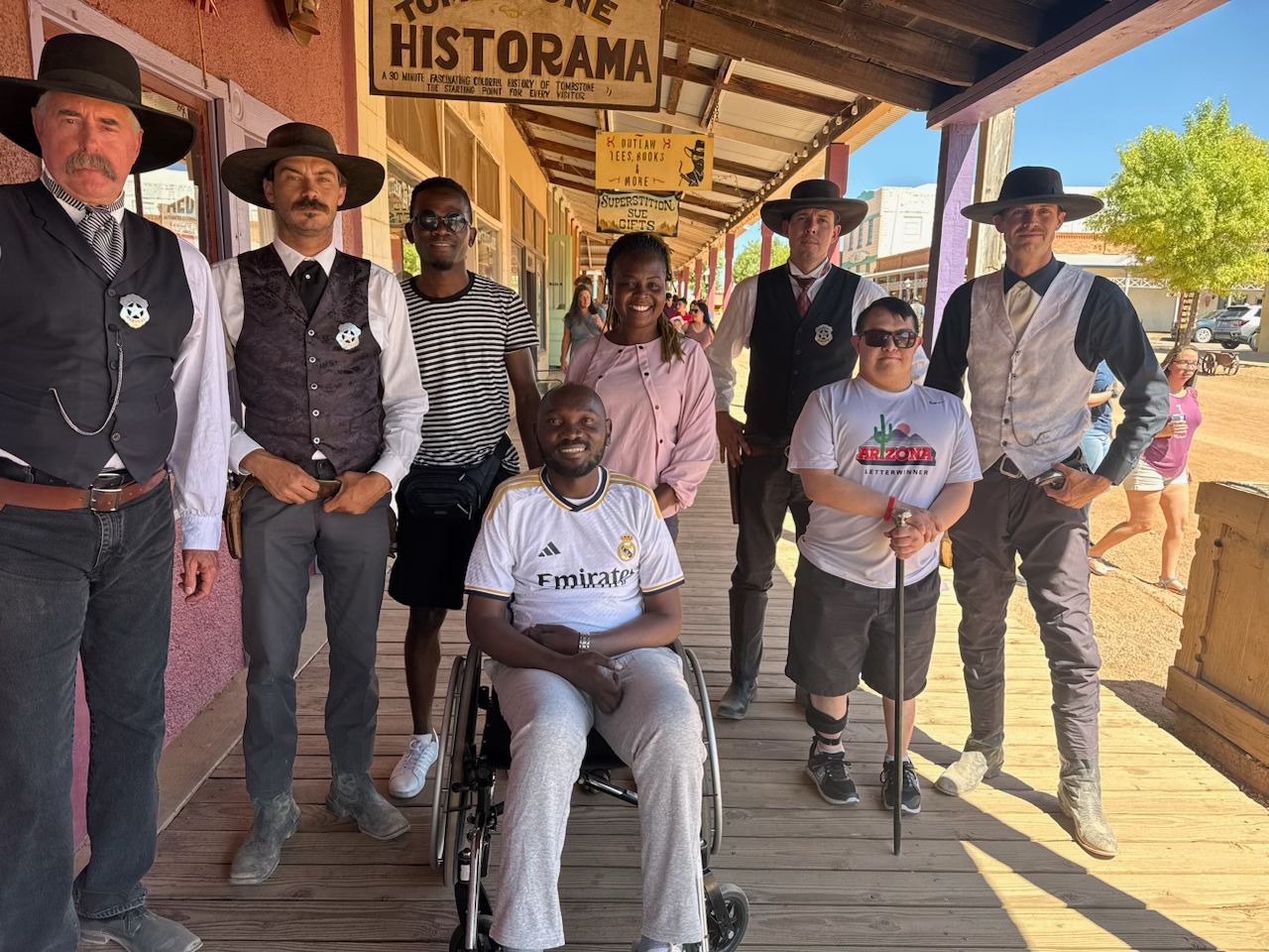 A group of eight people, seven men and one woman, posing for a photo and smiling at the camera. Everyone is standing except for one man in a wheelchair.    