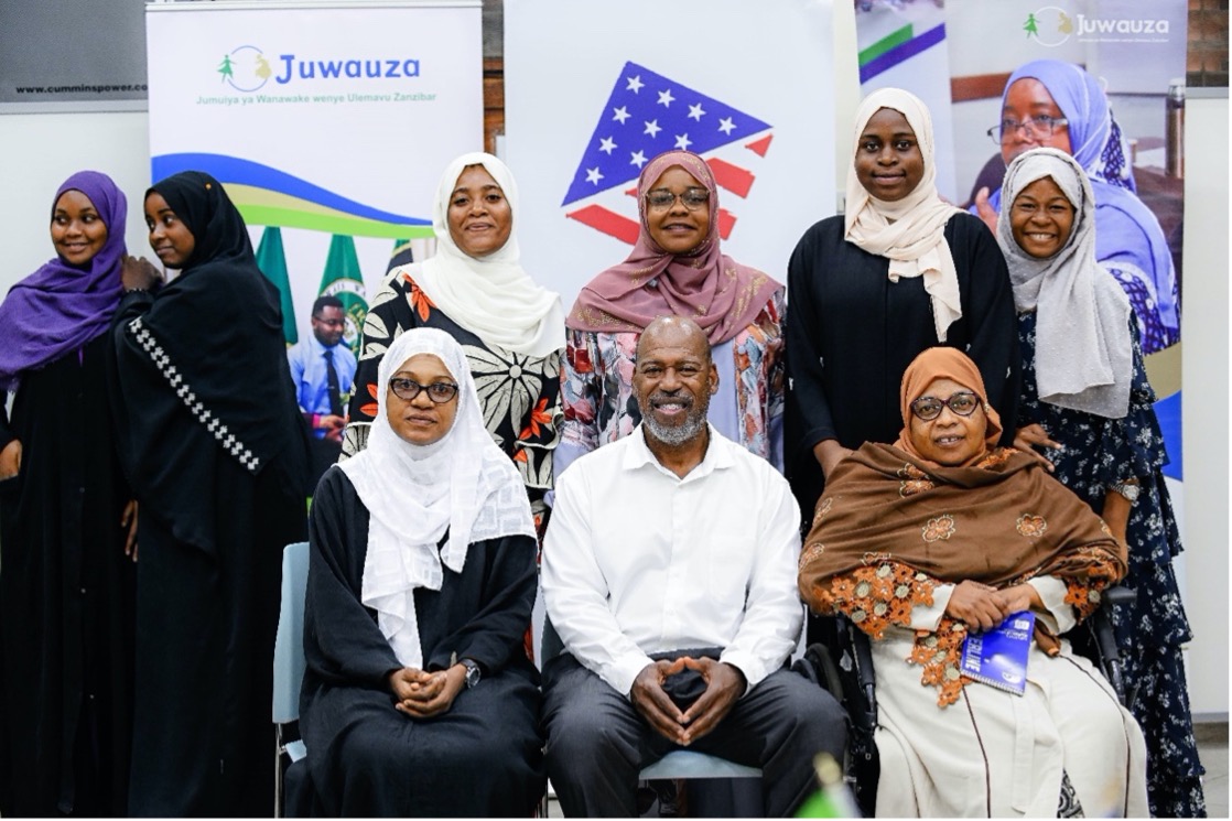 Nine individuals, including 8 Zanzibari women and one African American man, posing for a group photo. Behind them are several banners that read JUWAUZA.