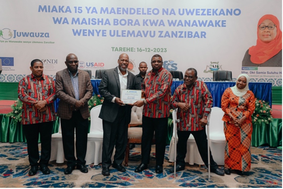Six people, including 5 Tanzanians and 1 American, posing for a group photo. The two individuals in the center of the group are holding up a certificate. 