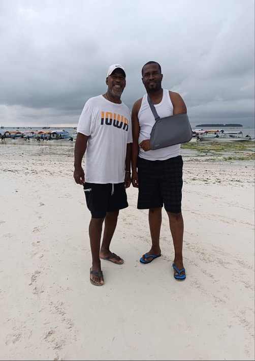 Two men, one African American man and one Zanzibari man with his arm in a sling, standing on a beach with boats behind them. 
