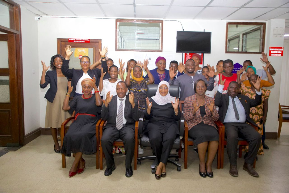 A group of 21 people, five of whom are seated in chairs, posing for a photo in an office building with their hands raised in the air. 