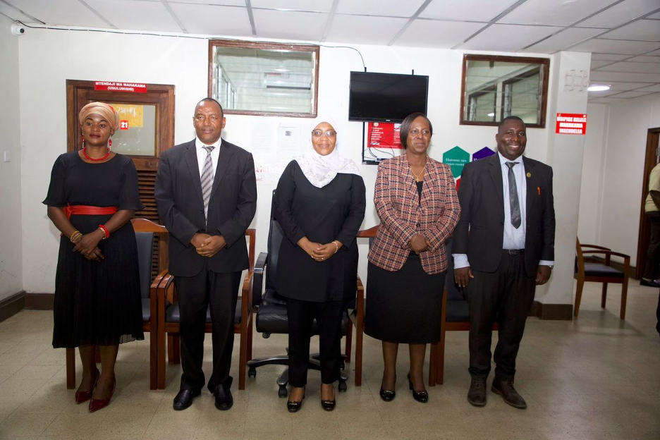 A group of five individuals, including two men and three women wearing professional workplace attire, pose for a group photo in an office building. 