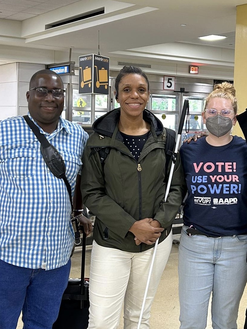 Three people, one African man, one African American woman, and an American woman, posing for a group photo, smiling into the camera. They seem to be at the airport. The Tanzanian man has short hair and is wearing glasses and a white and blue striped shirt with blue jeans. The African American woman has long black hair tied in the back. She is wearing a dotted blouse and a jacket with white trousers. She also has a white cane. The American woman has blond hair tied in the back, glasses, and a face mask. She is dressed in a black shirt with the wording “Vote! Use Your Power!” and light blue jeans.    