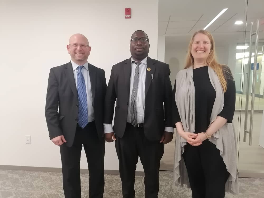 Three people, one American man, one Tanzanian man, and one American woman posing for a photo, smiling into the camera. The American man has short hair and a light beard, and is wearing a white shirt, blue tie, and grey suit. The Tanzanian man has short hair and is wearing glasses. He is dressed in a white shirt, grey tie, and a grey suit. The American woman has long blond hair and is dressed in a full black dress and a grey cardigan.