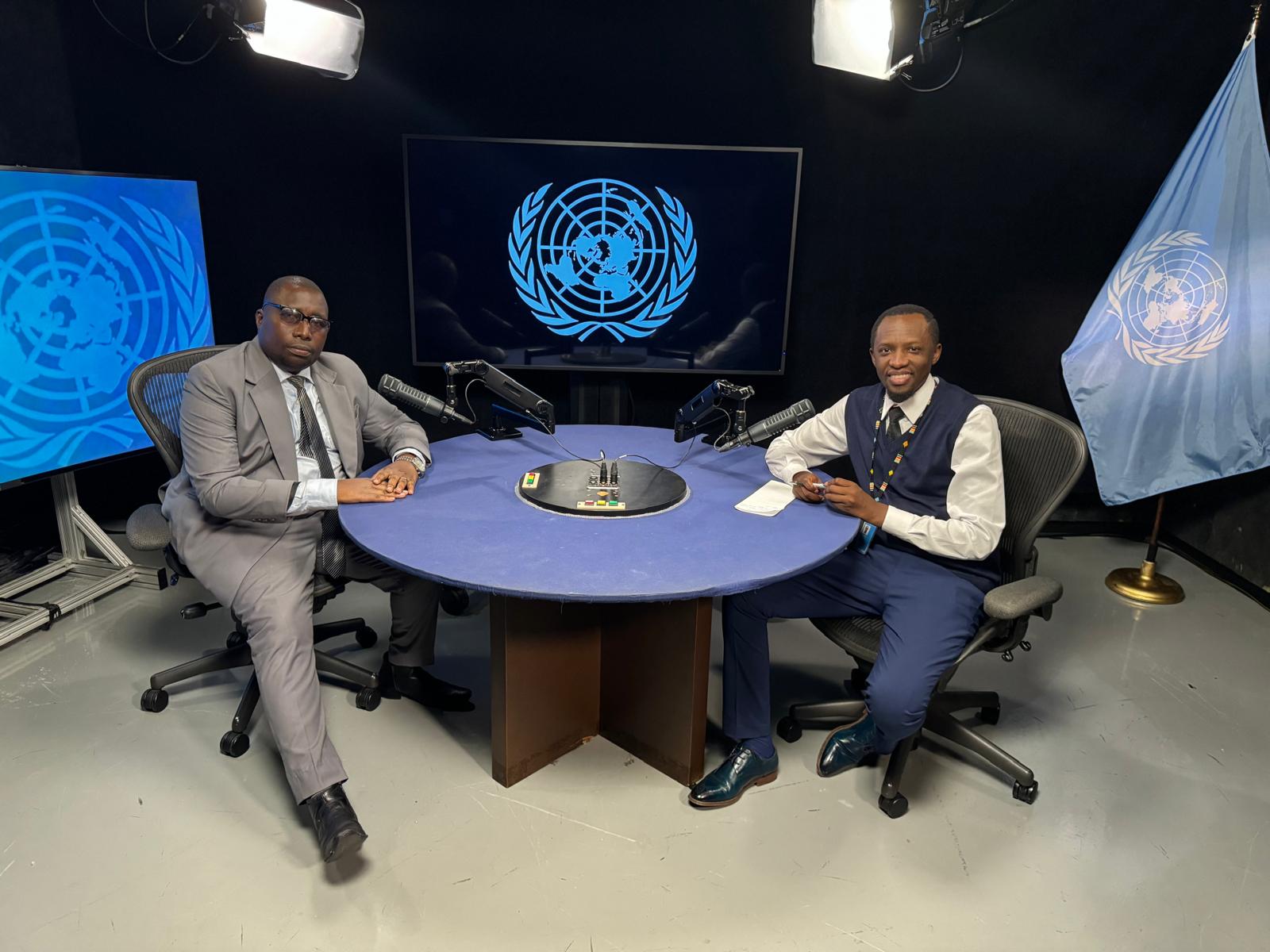 Two African men are sitting at a round table in a newsroom, smiling into the camera. The man on the left is wearing a white shirt with a tie and a grey suit. The man on the right is wearing a white shirt and tie with a blue vest and pants. In the background are two TV screens showing the UN logo. On the right side is a UN flag. 