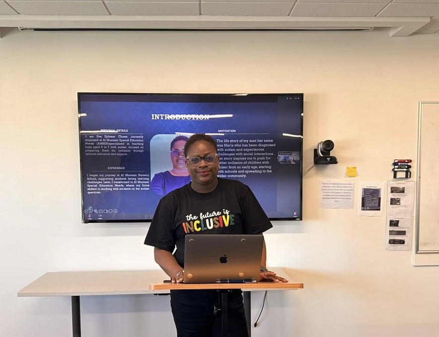 A Tanzanian woman with short hair, wearing glasses and a black T-shirt that reads "The Future is Inclusive," stands in front of a podium with a laptop in front of her. Behind her, a screen displays her image and text. The woman appears to be giving a presentation. 