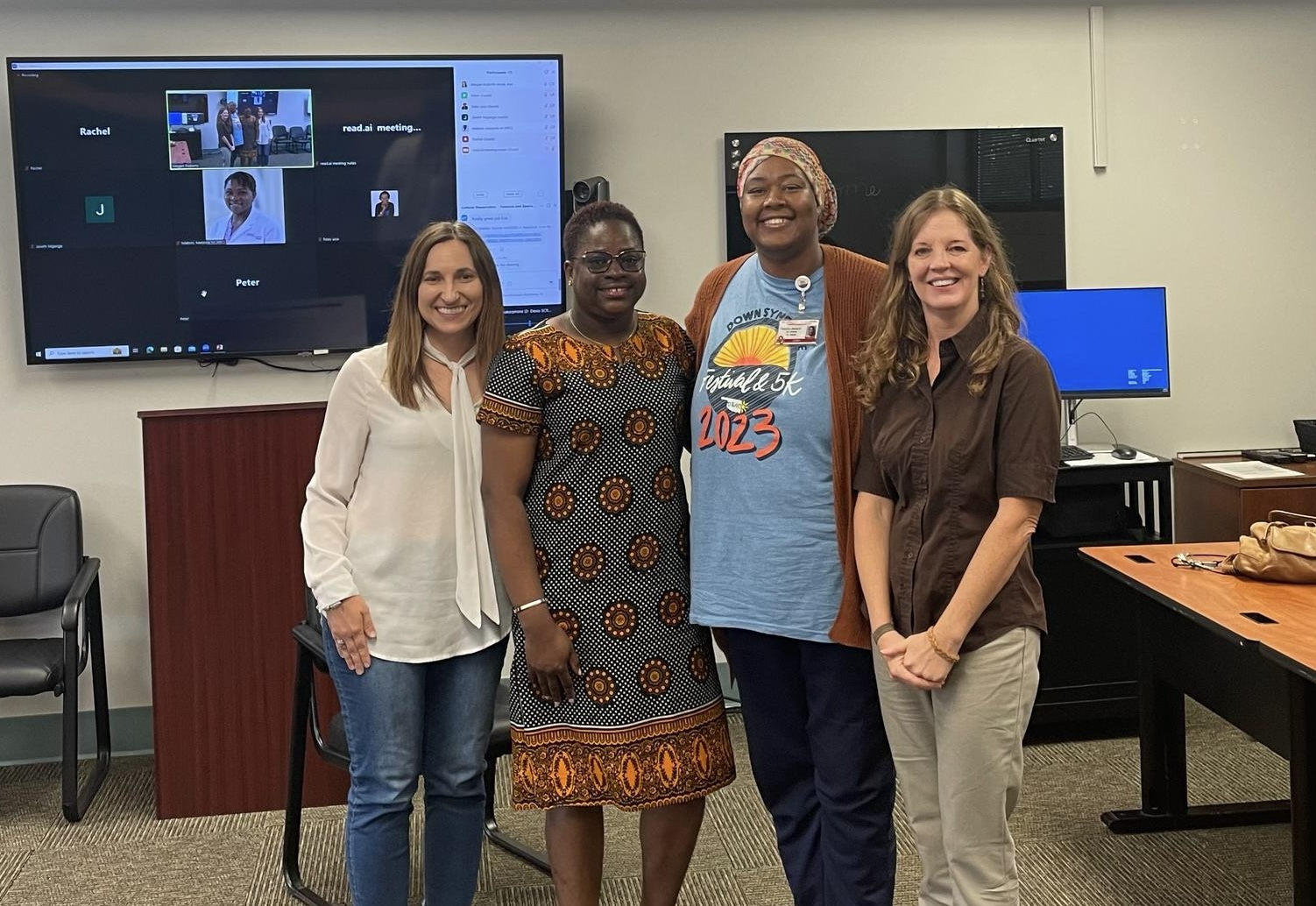 Four women posing for a group picture in a classroom, smiling at the camera. Three are American and one is Tanzanian. Behind them is a large screen, with chairs and tables in the background. The woman on the far left has long brown hair and is wearing a white blouse and blue jeans. The woman next to her has short black hair, dark-rimmed glasses, and is wearing an African print dress. The woman next to her is wearing a headscarf, a blue t-shirt with designs, a brown cardigan, and black pants. The woman on the far right has long brown hair and is wearing a brown blouse and grey pants. 