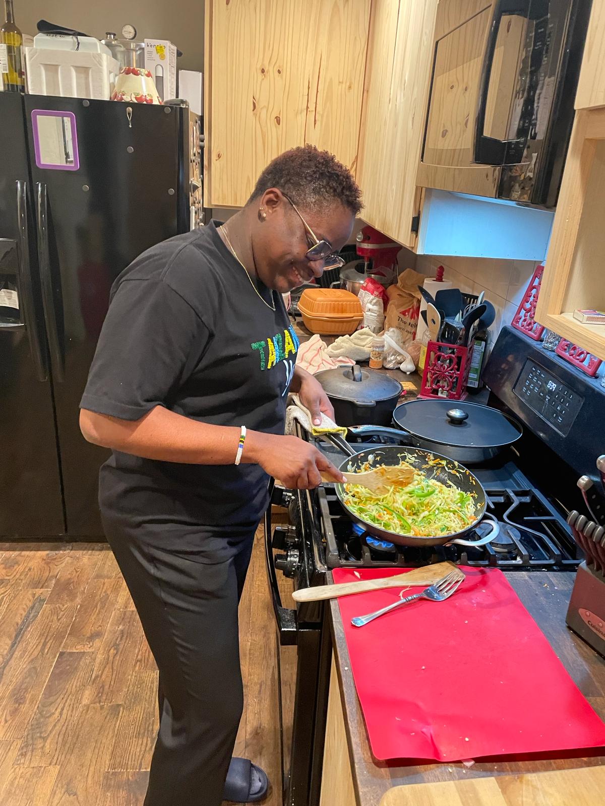 A Tanzanian woman dressed in a black t-shirt and pants, wearing dark-rimmed glasses, standing at a stove in a kitchen, cooking a meal. 