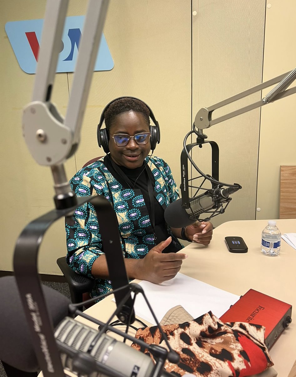 A Tanzanian woman seated at a table in a broadcasting studio. She is wearing headphones, an African print blouse, and glasses. There are two microphones in front of her, a bottle of water on the table, and a MiFi device. The wall behind her displays the "VOA" logo.