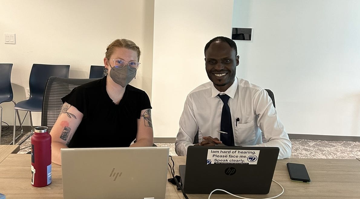 A white woman with long braided blond hair sits behind a laptop next to an African man where a button shirt and tie.