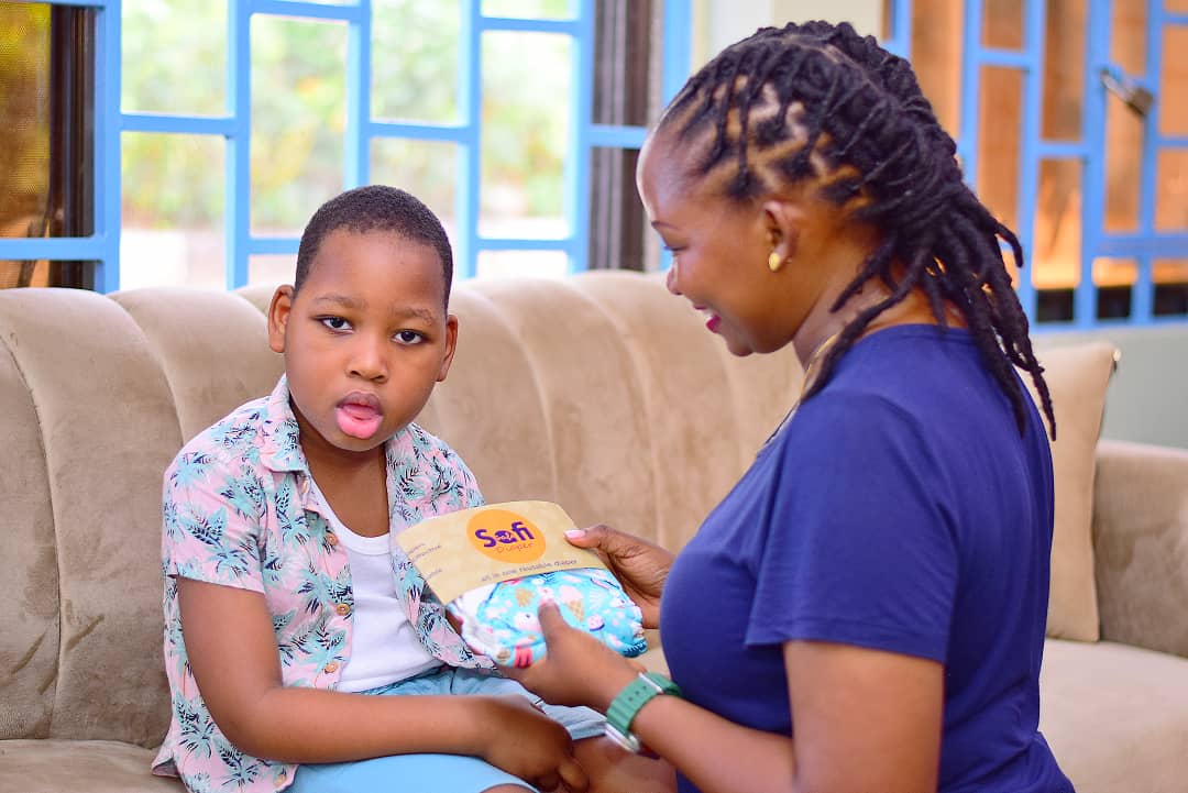 An African woman with braided hair holds a packaged diaper in front of a young African boy sitting on a couch. The boy is looking directly into the camera, while the women is facing him.  