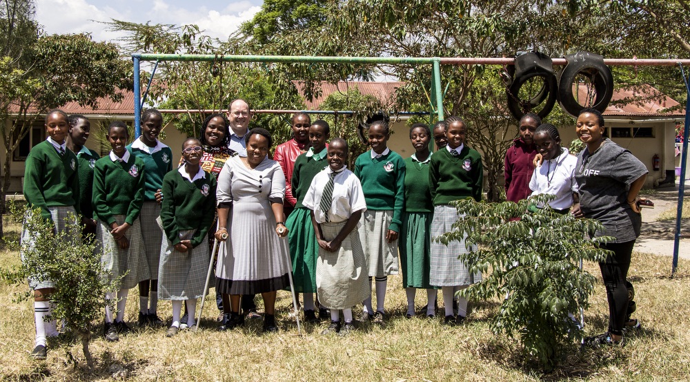 A large group of individuals stands in a yard, posing for the camera. One white American man is at the back of the group. Most of the group members, who are young Kenyan women, are wearing school uniform sweaters and skirts. One woman in the front of the group is standing with crutches.