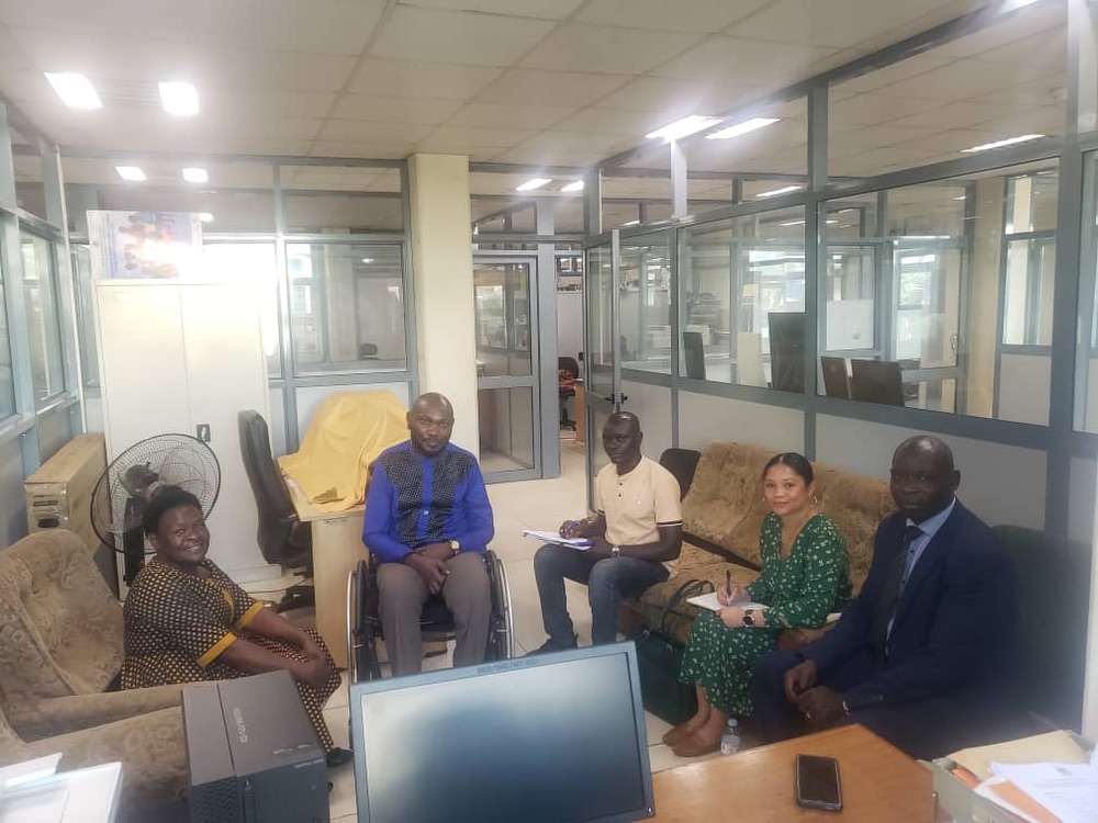 A group of people, including two women (one American and one Ugandan) and three men (both Ugandan), are meeting in an office space. One of the men is seated in a wheelchair.