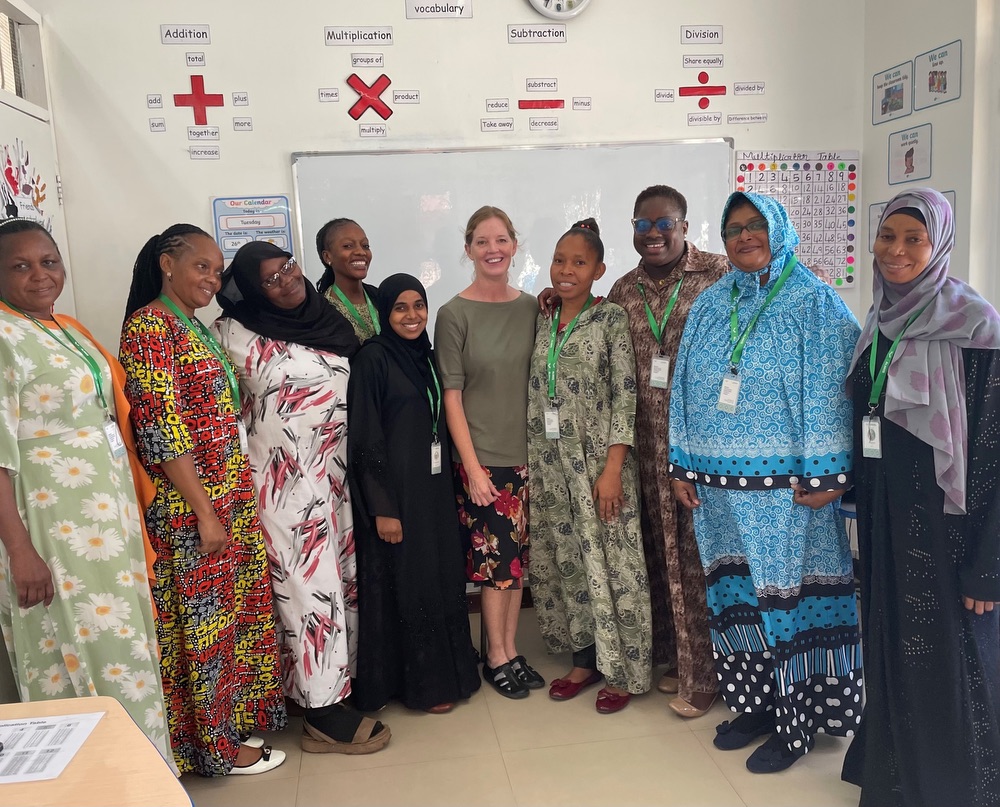 A group of 10 women, including one American woman (center) and 9 Tanzanian women, in a classroom, posing for a group picture. 