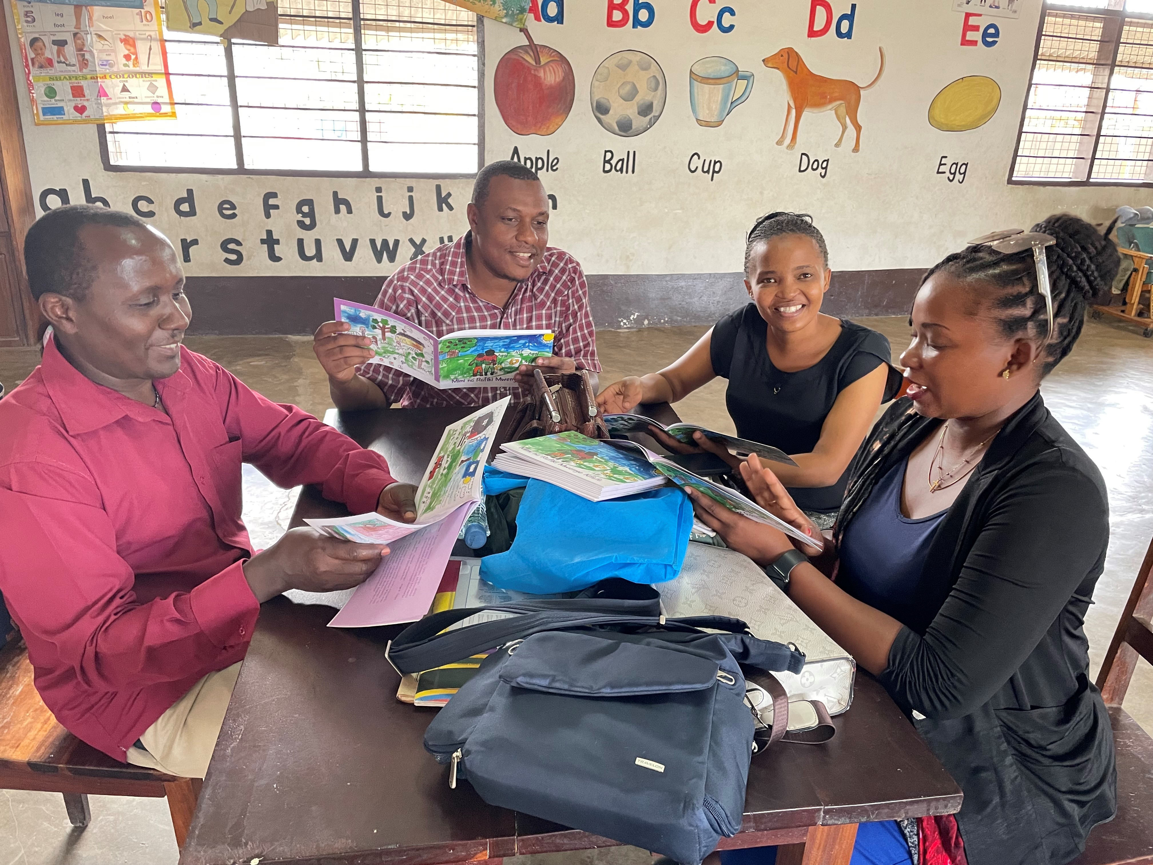 A group of Tanzanian people, two women and two men, sit at a table, each holding and looking at a picture book. 