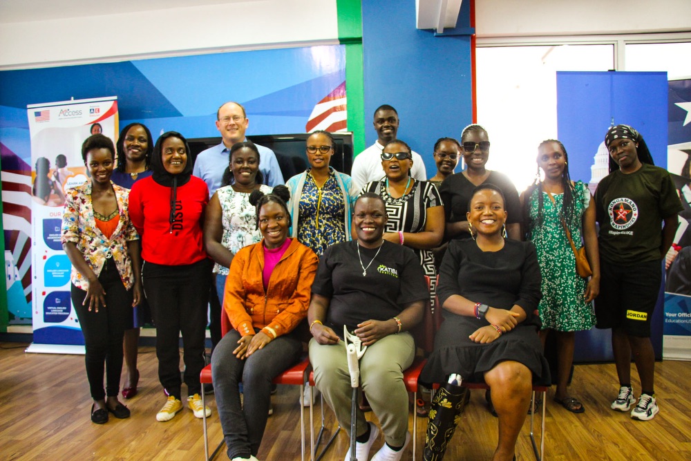 A group of individuals, including men and women, posing for a group photo in an indoor setting with banners and posters in the background. Some individuals are seated, while others stand behind them, all smiling at the camera. A few participants use mobility aids. The setting appears to be a professional or educational event.