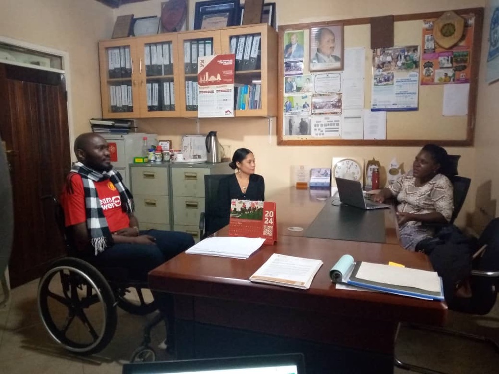 Three people, including two Ugandans (one man and one woman) and one American woman, meeting in an office space. The man is seated in a wheelchair.