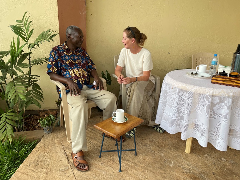 Two people, one older Tanzanian man (left) and one American woman (right), seated on chairs on a veranda, talking with each other.