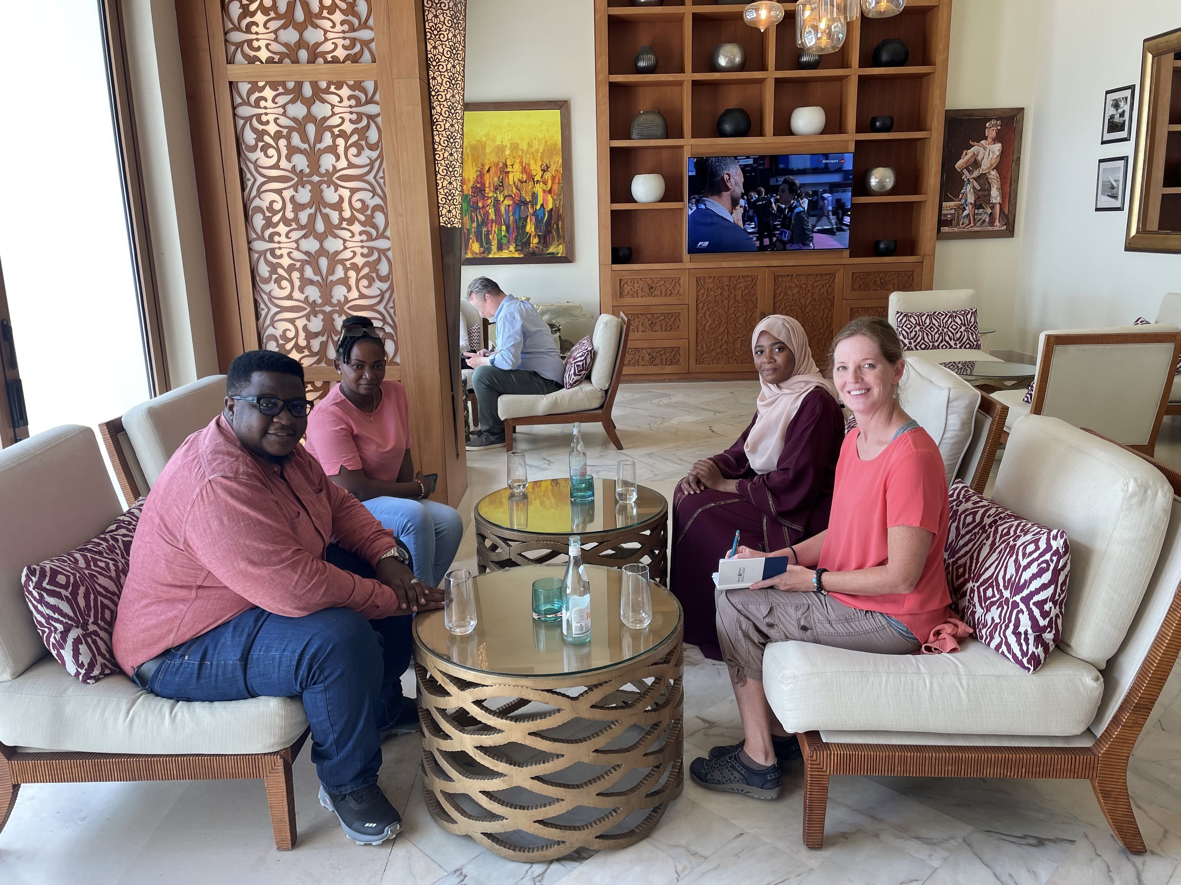 Four people, one Tanzanian man, two Tanzanian women, and one white, American woman with a notepad sit around coffee tables.